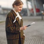 Train Ticket - Smiling female passenger wearing trendy plaid coat and white blouse checking passport and ticket standing on pavement near contemporary building of airport