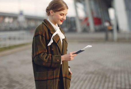 Train Ticket - Smiling female passenger wearing trendy plaid coat and white blouse checking passport and ticket standing on pavement near contemporary building of airport