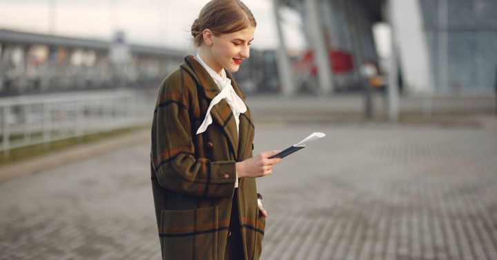 Train Ticket - Smiling female passenger wearing trendy plaid coat and white blouse checking passport and ticket standing on pavement near contemporary building of airport