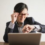 Delays - Young frowning man in suit and glasses looking at wristwatch while waiting for appointment sitting at desk with laptop
