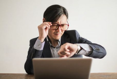 Delays - Young frowning man in suit and glasses looking at wristwatch while waiting for appointment sitting at desk with laptop