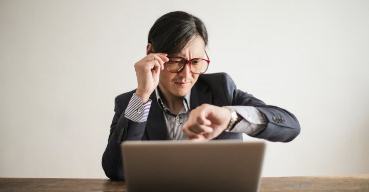 Delays - Young frowning man in suit and glasses looking at wristwatch while waiting for appointment sitting at desk with laptop