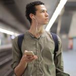 Train Apps - Young ethnic man in earbuds listening to music while waiting for transport at contemporary subway station