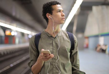 Train Apps - Young ethnic man in earbuds listening to music while waiting for transport at contemporary subway station