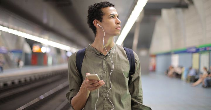 Train Apps - Young ethnic man in earbuds listening to music while waiting for transport at contemporary subway station