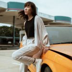 Quiet Cars - Slender young Asian female in casual wear looking away while standing leaned on one leg near old fashioned car parked on asphalt road on windy weather under cloudy sky