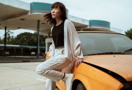 Quiet Cars - Slender young Asian female in casual wear looking away while standing leaned on one leg near old fashioned car parked on asphalt road on windy weather under cloudy sky