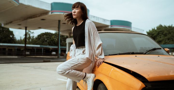 Quiet Cars - Slender young Asian female in casual wear looking away while standing leaned on one leg near old fashioned car parked on asphalt road on windy weather under cloudy sky